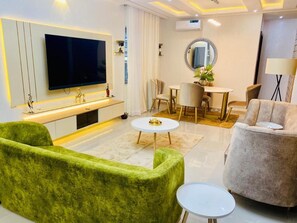 A modern living room with a flat-screen TV, green and beige sofas, a round coffee table, and a dining area with a white table and chairs. - Wonder Court 2 Hotel and Apartment (Lekki)