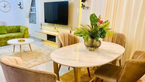A modern living room with a round table, beige chairs, a green sofa, a flat-screen TV, and a decorative plant.