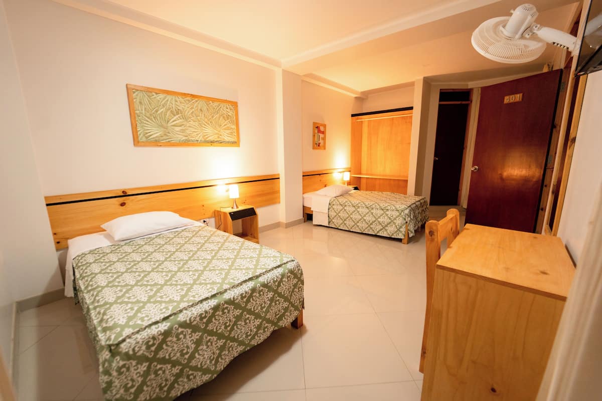 Quarto de hotel com duas camas, uma escrivaninha de madeira, uma cadeira, um ventilador e uma porta com a inscrição '401'.