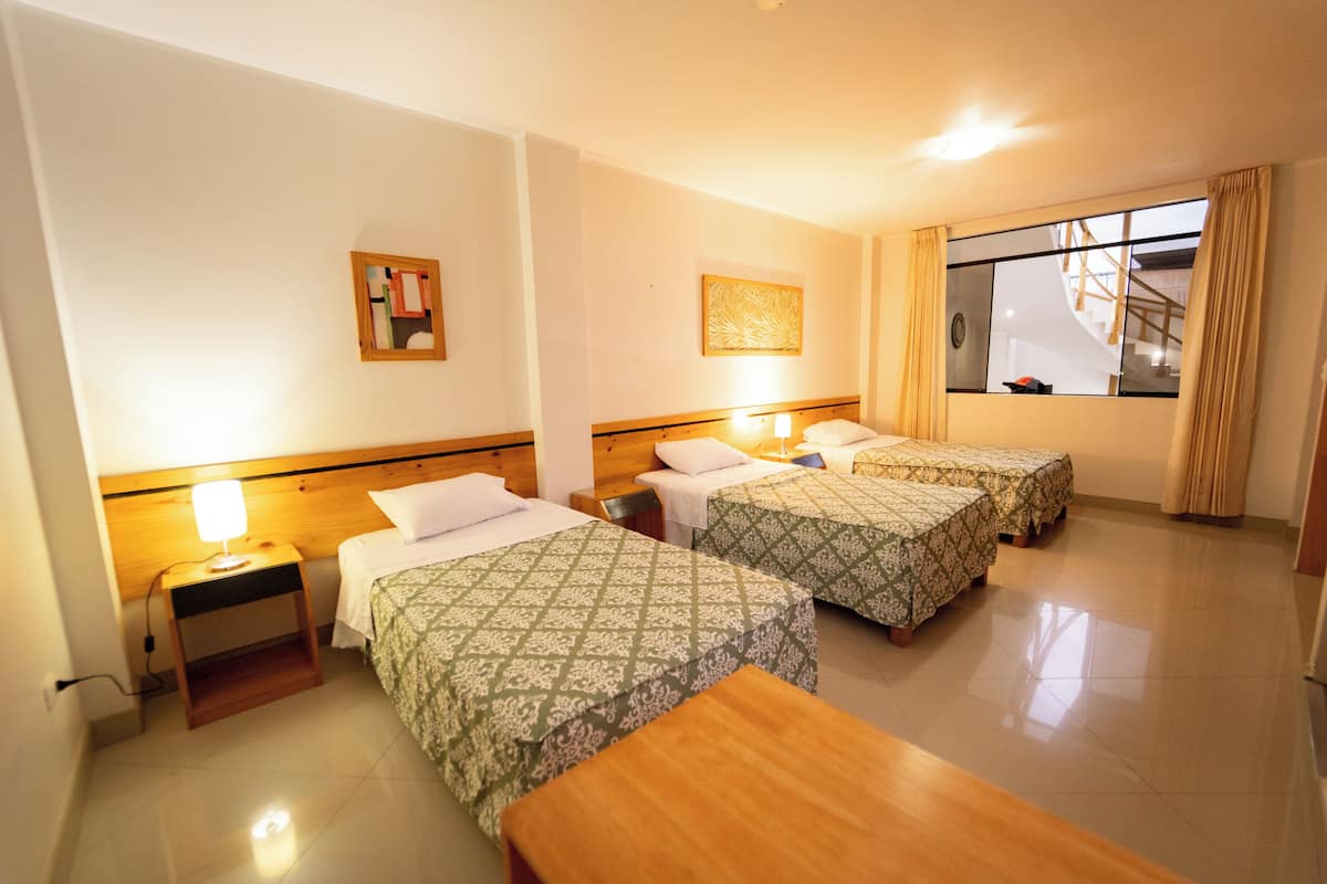 Quarto de hotel com duas camas de solteiro, uma mesa de cabeceira de madeira, uma pequena mesa e uma janela com cortinas.