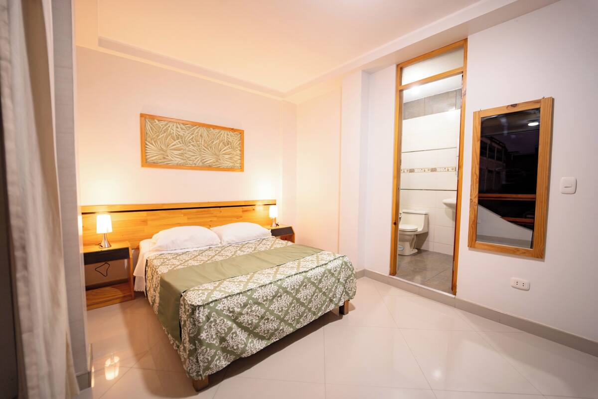 Quarto de hotel com cama, luminárias de cabeceira, obra de arte pendurada na parede, espelho e banheiro visível através de uma porta aberta.