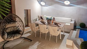 Uma área externa moderna para refeições, com uma mesa redonda e cadeiras, uma cadeira suspensa, um banco branco e uma piscina pequena.