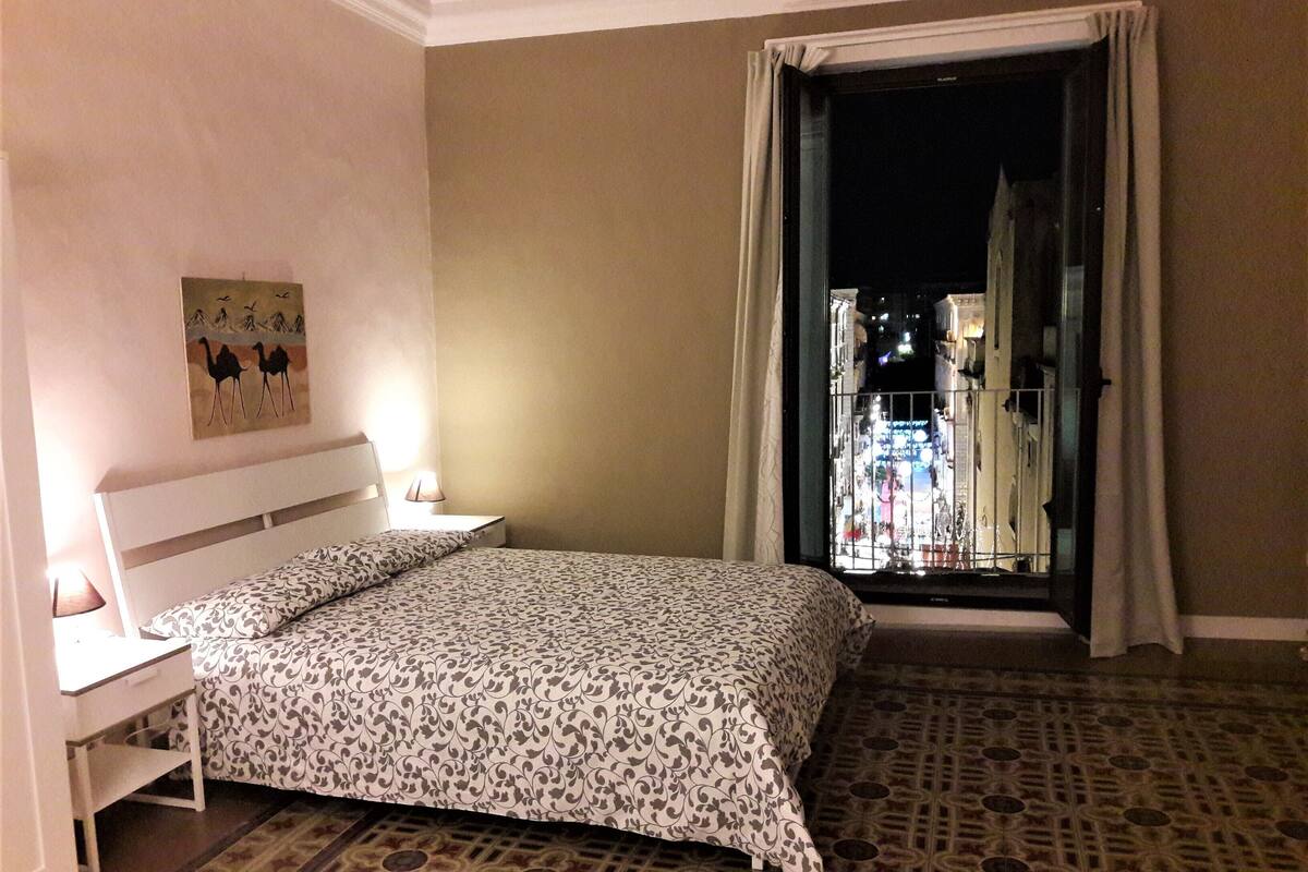 Ein Hotelzimmer mit Bett, Nachttisch, Lampe und Blick auf eine Stadtlandschaft durch eine geöffnete Tür.
