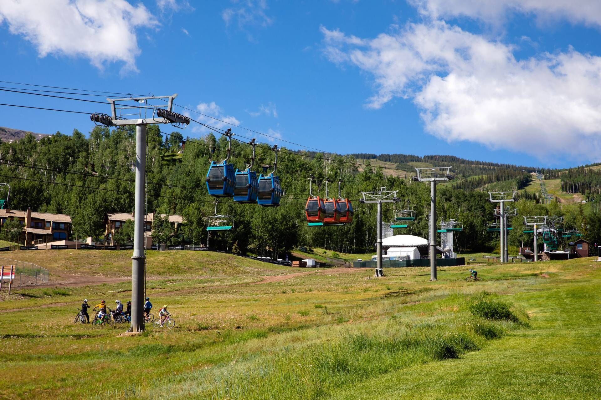 A ski lift with blue and red chairs, a grassy area with cyclists, and a mountainous backdrop.