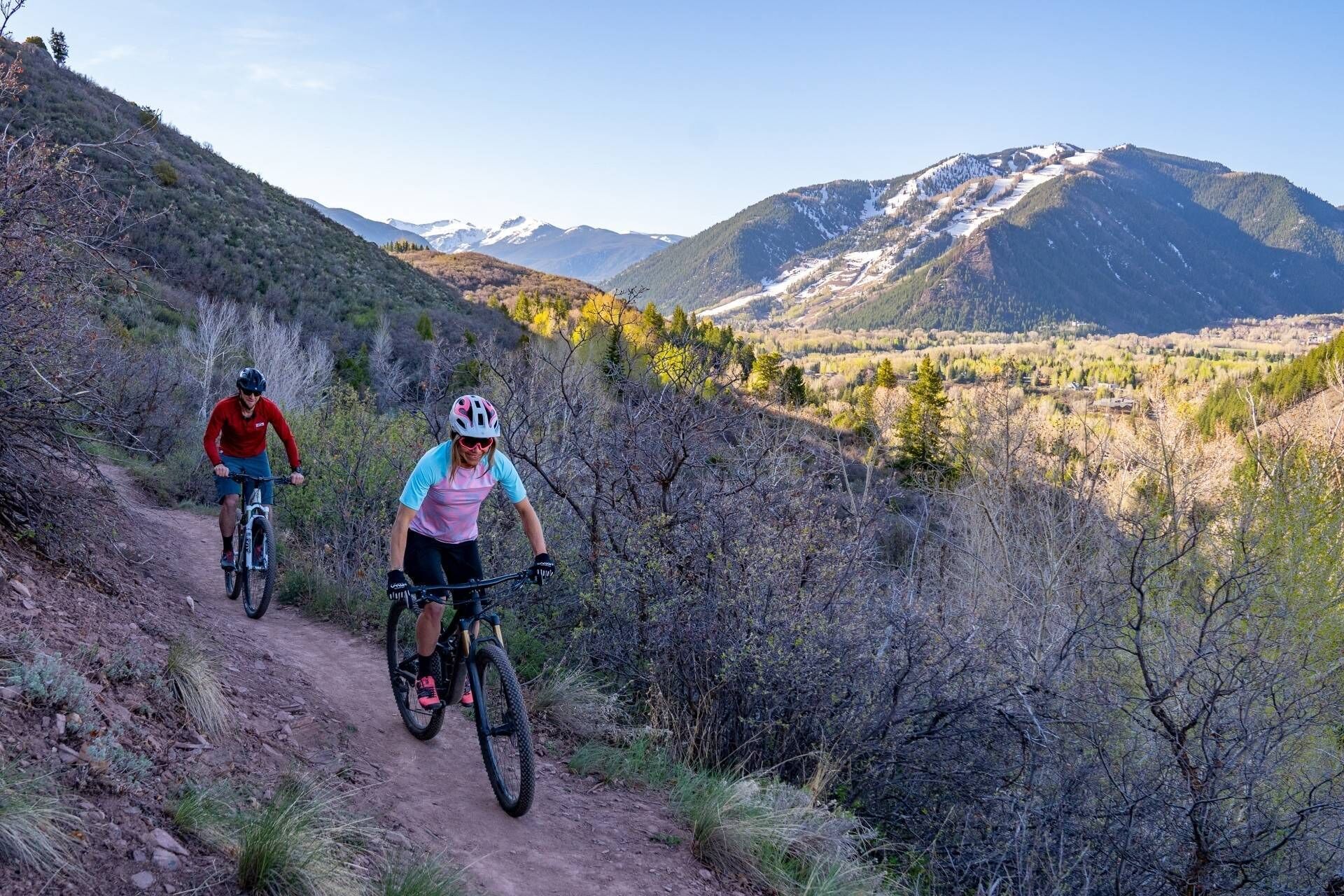 Two cyclists on a dirt trail with mountainous terrain and sparse vegetation in the background.