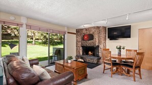 A living room with a stone fireplace, a TV mounted on the wall, and a dining area with wooden furniture. - Lichenhearth 08 (Snowmass Village)