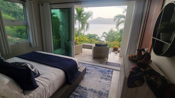 A bedroom with a large bed, a sliding glass door leading to a balcony with a view of the sea and mountains. - BEACHFRONT 5 BEDROOM VILLA–POOL, SOLAR, PRIVATE (East End)