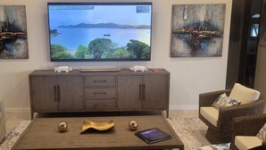 A living room with a TV displaying a scenic view, a wooden coffee table with decorative items, a sofa, and a wicker armchair. - BEACHFRONT 5 BEDROOM VILLA–POOL, SOLAR, PRIVATE (East End)