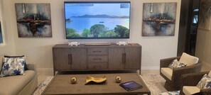 Een woonkamer met een televisie waarop een mooi landschap te zien is, een houten salontafel met decoratieve objecten, een bank en een rieten fauteuil.