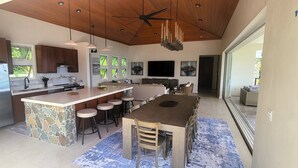 A modern kitchen with a central island, bar stools, a dining area, and a large TV. - BEACHFRONT 5 BEDROOM VILLA–POOL, SOLAR, PRIVATE (East End)