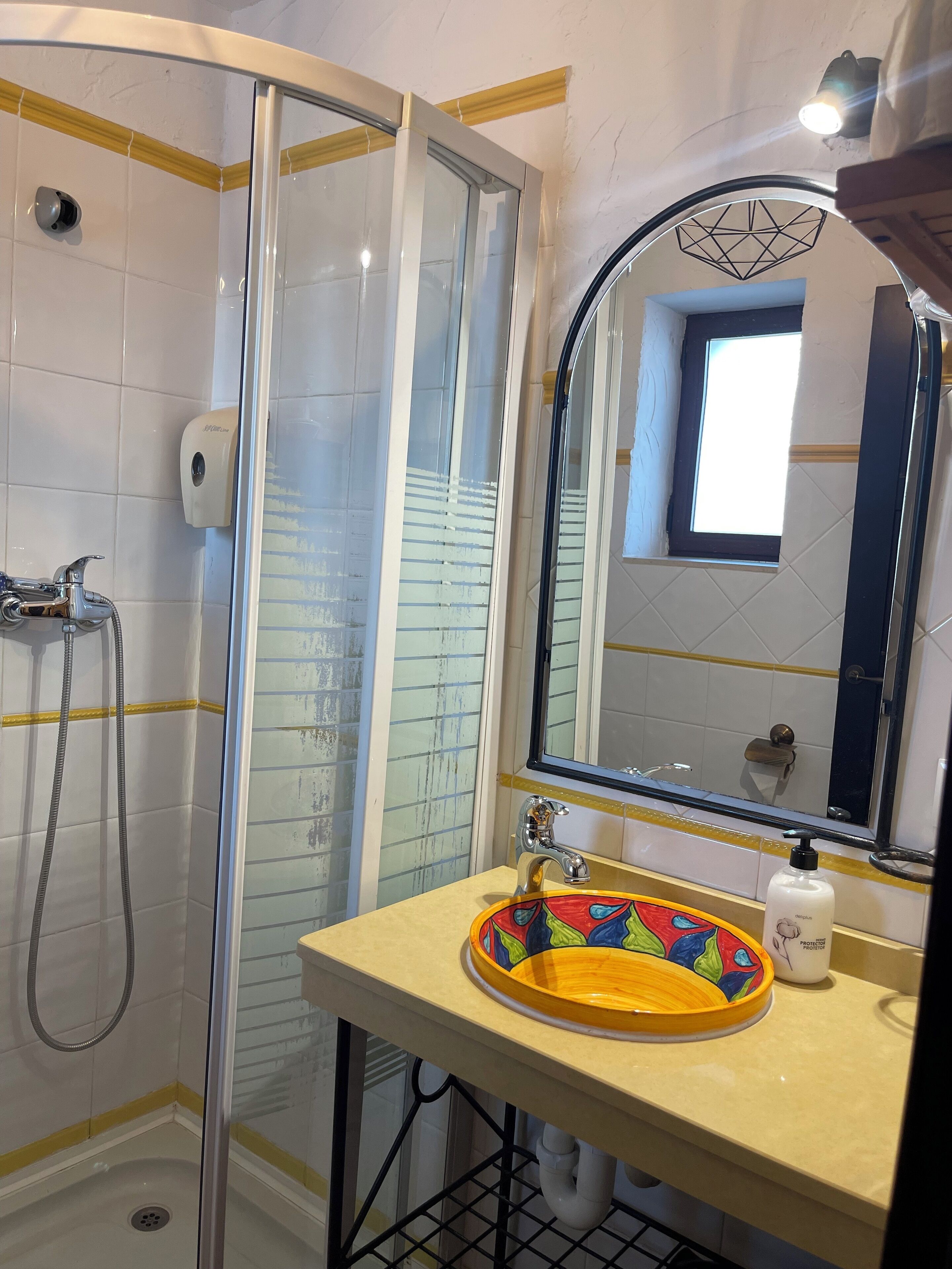 Une salle de bain dotée d’une douche à parois de verre, d’un lavabo avec un récipient aux couleurs vives et d’un miroir orné d’un motif géométrique au-dessus.