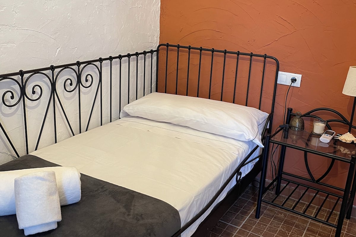 Una cama individual con estructura metálica, ropa de cama blanca y cabecera negra. Hay una mesita de noche pequeña con una lámpara y un vaso de leche. Las paredes están pintadas de blanco y naranja.