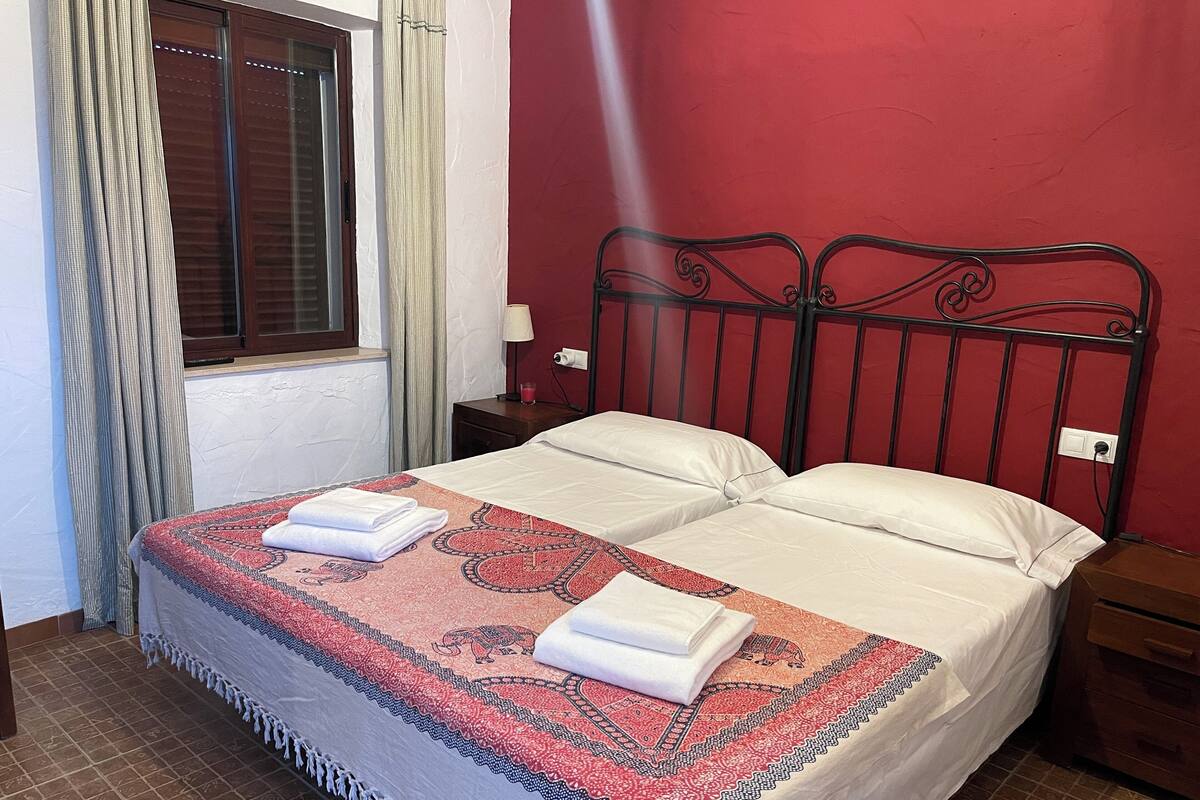 Cama doble con una colcha estampada y dos toallas dobladas, una mesita de noche con lámpara, y una ventana con cortinas.