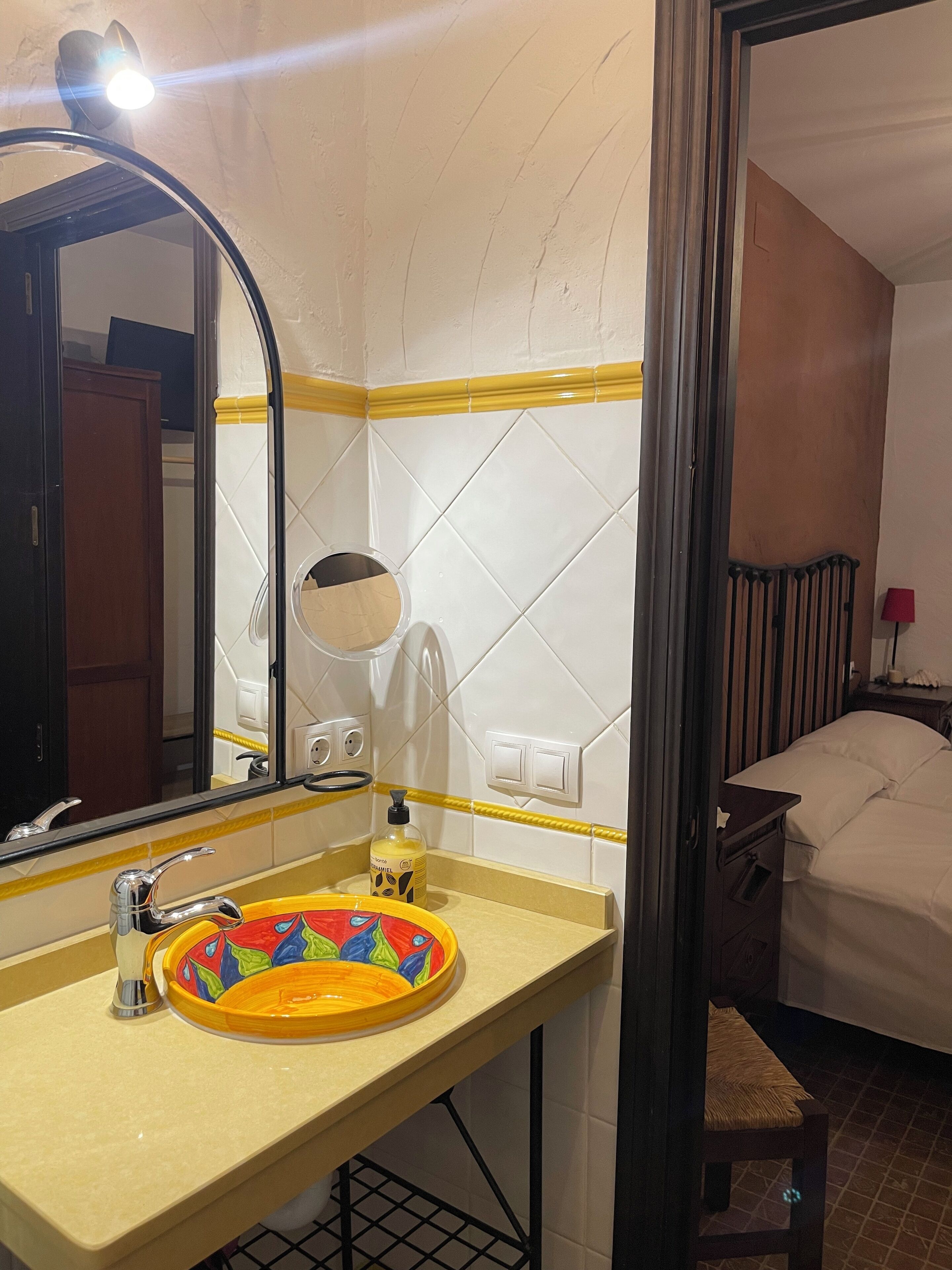 Ein Badezimmer mit Waschbecken, Spiegel und einem durch eine geöffnete Tür erkennbaren Bett.