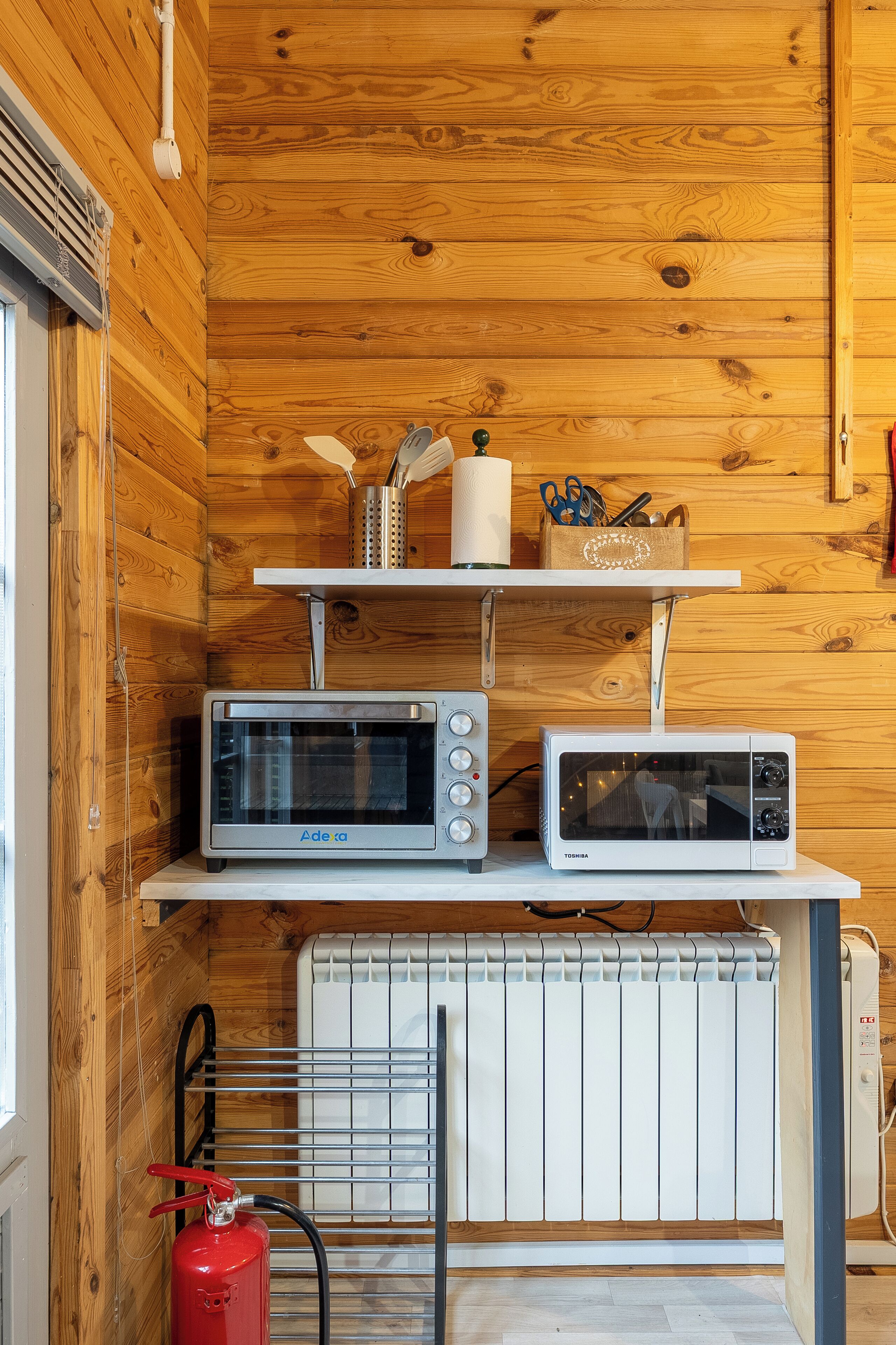 A kitchen with wooden walls, a white radiator, and two microwaves on a shelf.
