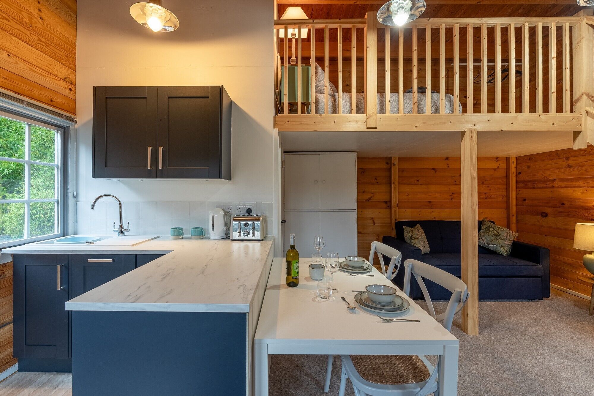 A modern kitchen with a central island, a dining area with a table set for two, and a loft bedroom with a wooden railing.