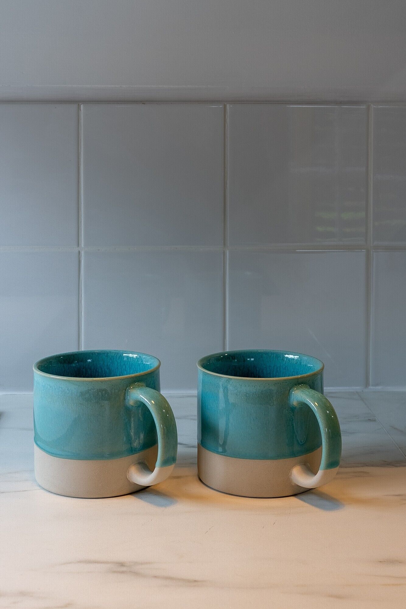 Two teal ceramic mugs with beige bases on a light-colored countertop against a tiled backsplash.