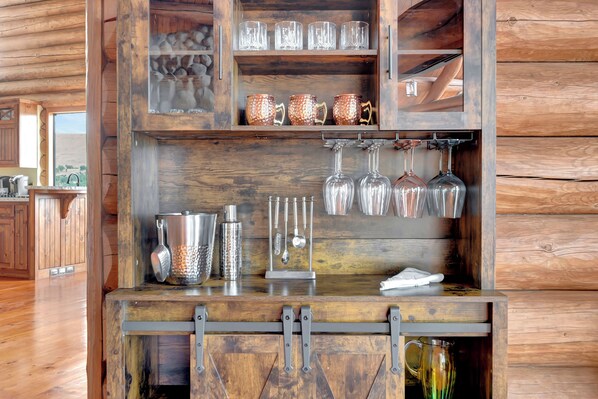 A wooden bar area with shelves holding glasses, mugs, and a grater.