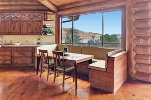A cabin-style kitchen with wooden cabinets, a dining table with chairs, a built-in bench, and a large window offering a view of the outdoors.