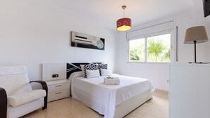 Un dormitorio moderno con una cama, mesitas de noche, una silla, una lámpara y una ventana con vistas a palmeras.
