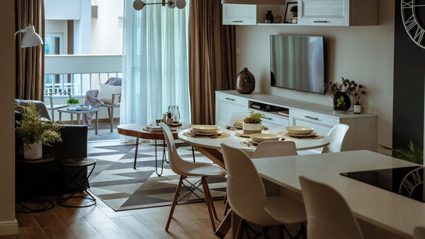 Et moderne spiseområde med et hvidt bord dækket til fire personer, en sofa og et lille sofabord med en plante. Et stort vindue med gardiner er synligt, og et fjernsyn er monteret på væggen over køkkenbordet.