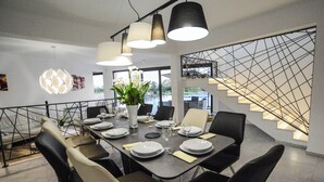 Ein moderner Essbereich mit einem rechteckigen Tisch, gedeckt für sechs Personen, umgeben von schwarzen und beige farbenen Stühlen. Über dem Tisch befinden sich hängende Pendelleuchten, und im Hintergrund ist eine Treppe mit einem geometrischen Geländer zu sehen.