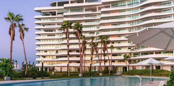 A multi-story hotel with a pool area, palm trees, and outdoor umbrellas. - Exclusive Acapulco Apt With Beach Access (Acapulco)