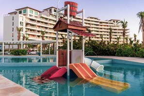 A swimming pool with slides and a play structure, surrounded by palm trees and a hotel building in the background. - Exclusive Acapulco Apt With Beach Access (Acapulco)