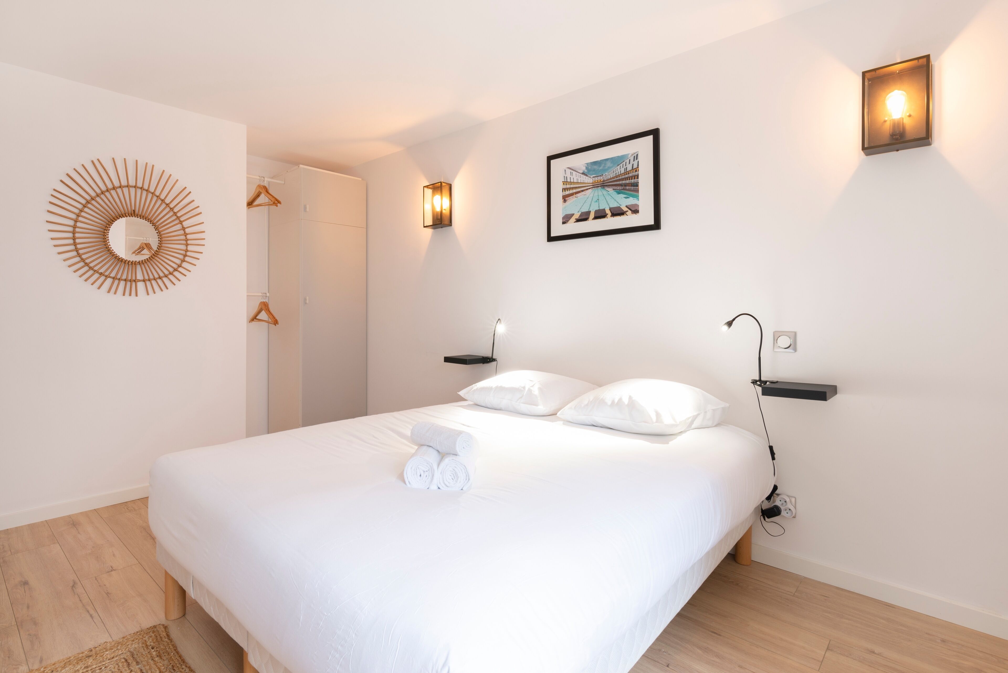 Un lit bien fait, avec du linge de lit blanc et des serviettes pliées, un grand miroir rond fixé au mur, un tableau encadré et deux lampes de lecture fixées au mur.
