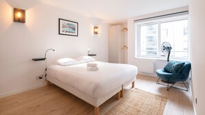 A modern bedroom with a large bed, bedside lamps, a framed picture, a window with a view, and a blue chair. - Le Spacieux de Dizier - Proche place des Vosges (Nancy)