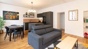 A modern living room with a grey sofa, a wooden dining table, and a kitchen area with dark cabinets and a built-in oven.