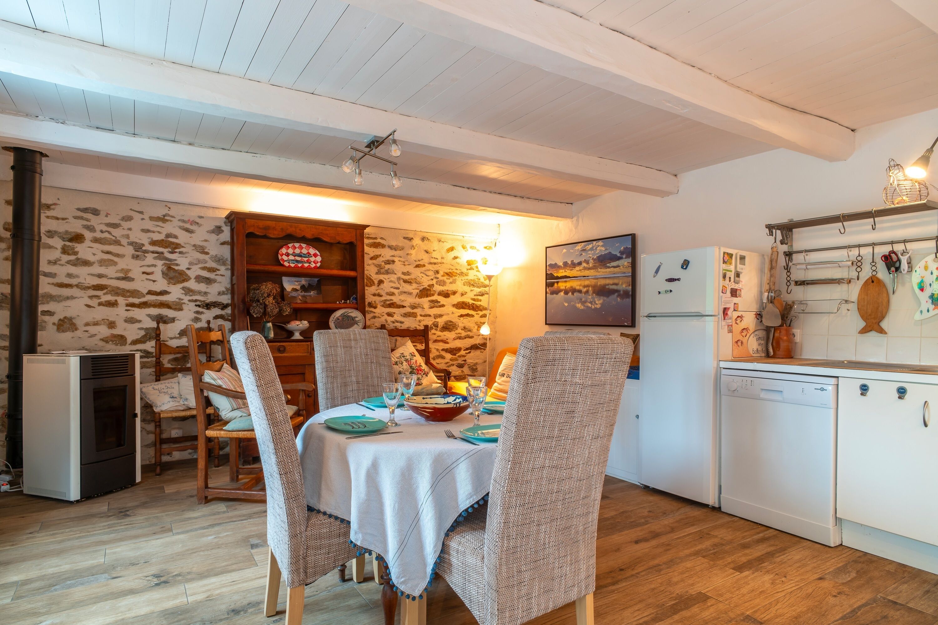 A cozy dining area with a stone wall, wooden chairs, a table set for two, and a kitchen with a refrigerator and dishwasher.