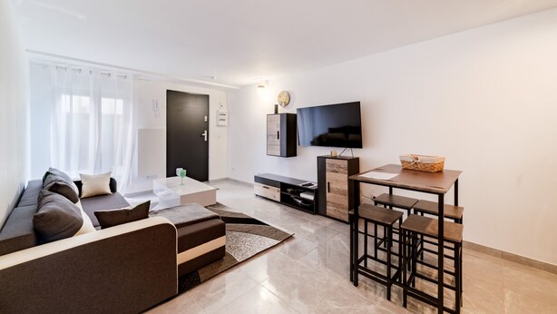 A modern living room with a sofa, a coffee table, a TV, and a small dining area with stools. - Batié - Appt 4 personnes à Mulhouse (Mulhouse)