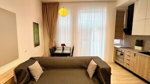 A compact living space with a sofa, a dining table with chairs, a kitchen area, and a large window with curtains. - NIce Flat in Villa Near Old Town
 (București)