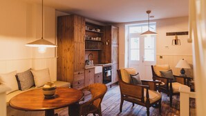 A cozy living room with a wooden dining table, wicker chairs, and a sofa. A kitchen with wooden cabinets and a window is visible in the background. - Rockpool Cottage 5 Buckingham Mews Coastal Retreat (Broadstairs)