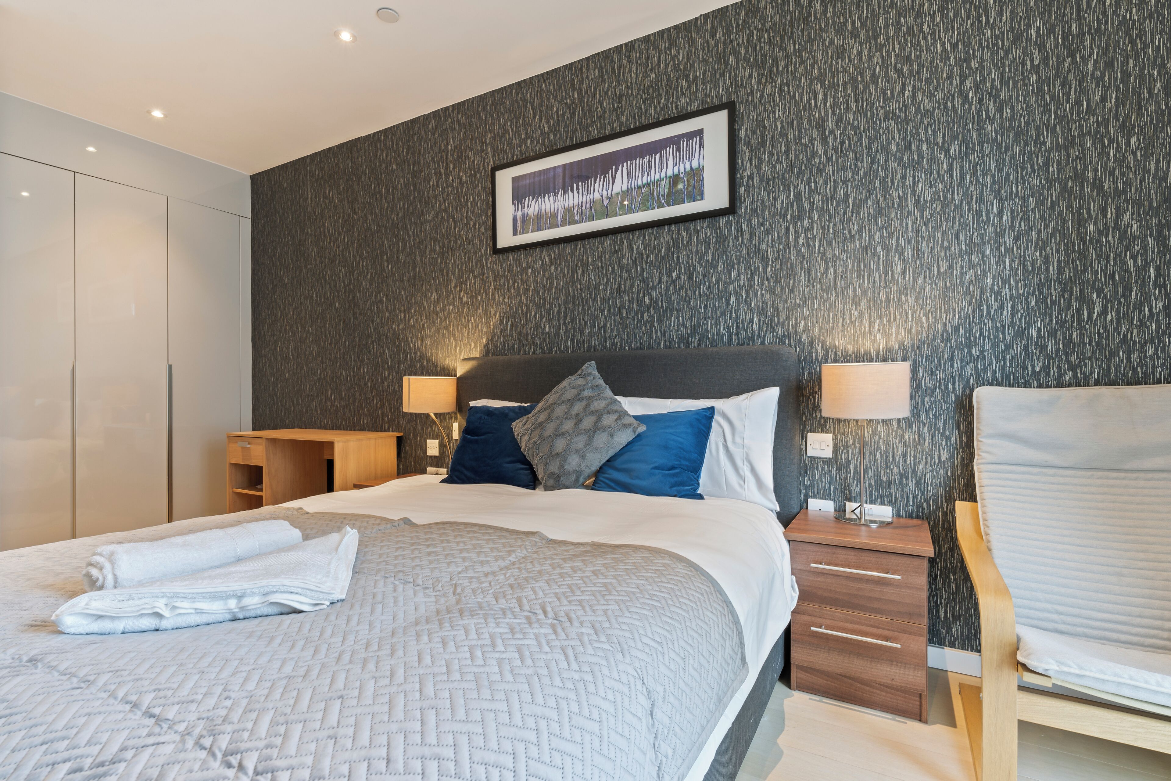 A modern hotel room with a large bed, a wooden nightstand, a desk, and a chair.