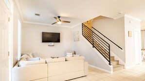 A living room with a beige sofa, a flat-screen TV mounted on the wall, a ceiling fan, and a staircase with black railings.