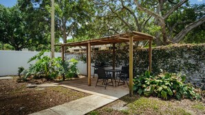 Un patio cubierto con una mesa y sillas, rodeado de plantas y una valla blanca.