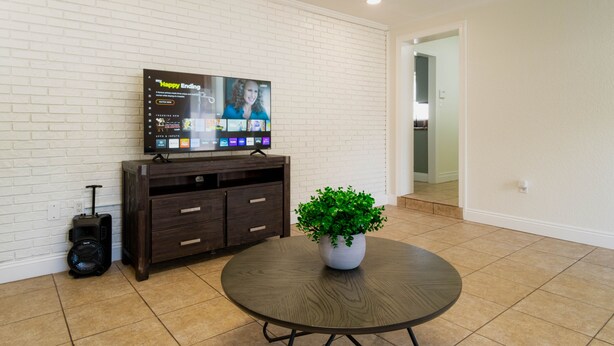 Una sala de estar con un televisor de pantalla plana en una pared de ladrillo blanco, un mueble de madera oscura, una mesa redonda con una planta en maceta y una maleta.