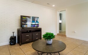 A living room with a flat-screen TV on a white brick wall, a dark wooden cabinet, a round table with a potted plant, and a suitcase. - Comfy house walk to Stadium (Tampa)