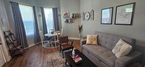 Un salon avec un canapé gris, une table basse, une table à manger avec des chaises et une fenêtre avec des rideaux. On peut y voir des horloges murales et des tableaux encadrés accrochés au mur.