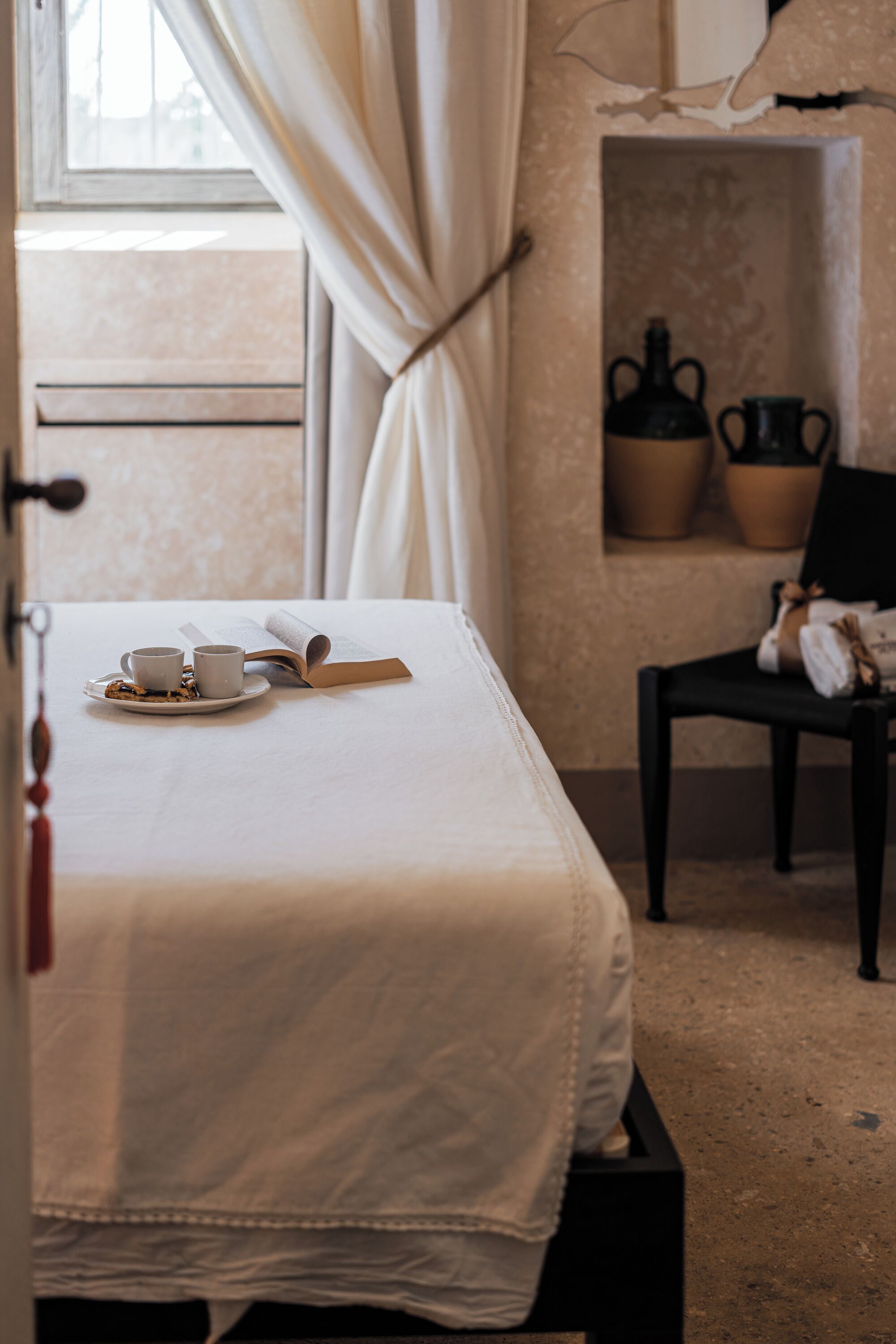 A neatly made bed with a tray of cookies and two cups, a chair, and a shelf with decorative items.
