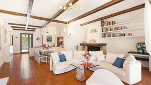 A spacious living room with a white sofa, a glass coffee table, and a fireplace. There is a bookshelf filled with books and a dining area with a table and chairs. - Villa Castiglione 9, Emma Villas (Castiglione della Pescaia)