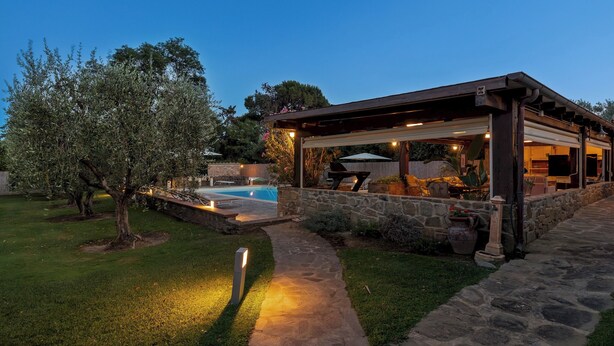 A covered outdoor seating area with stone walls and a pool in the background. - Villa Castiglione 9, Emma Villas (Castiglione della Pescaia)