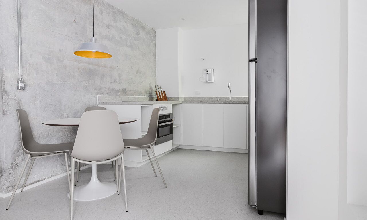 A modern kitchen with white cabinetry, a grey countertop, and a stainless steel refrigerator. There is a dining area with a white table and two grey chairs.