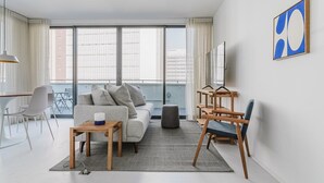 Ein modernes Wohnzimmer mit einer Couch, einem Couchtisch und Sesseln. Ein großes Fenster mit Jalousien, ein Balkon und eine Aussicht auf Gebäude.