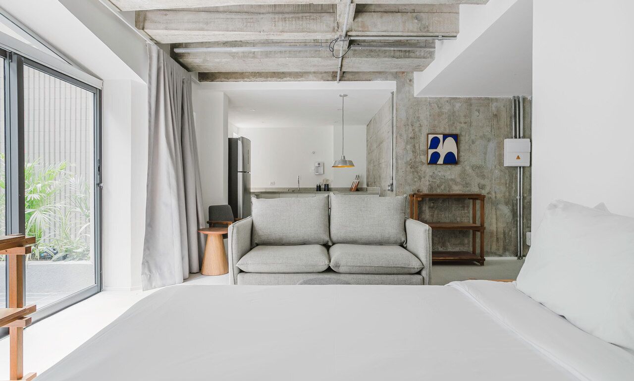 A modern hotel room with a large bed, a grey sofa, a small wooden side table, and a view of the outdoors through a large window.