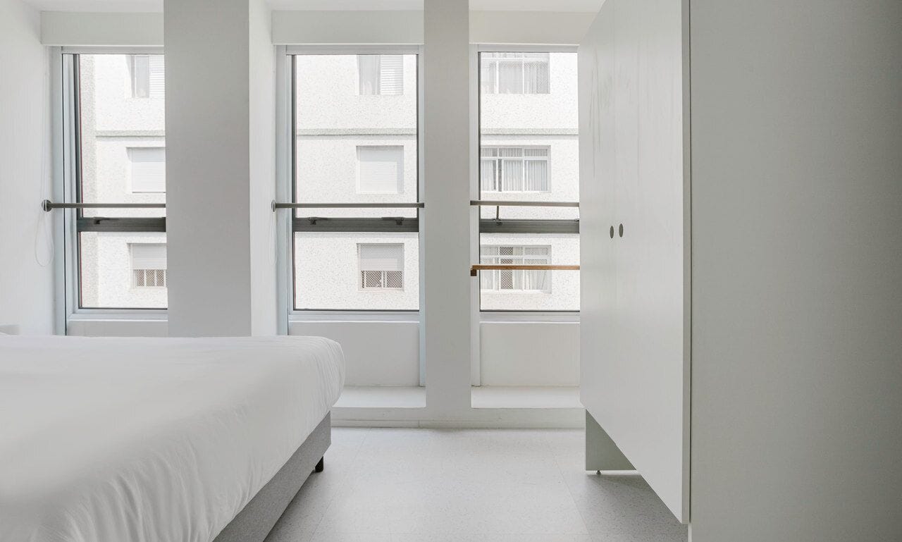 A minimalist bedroom with a large bed, a wardrobe, and a view of neighboring buildings through large windows.