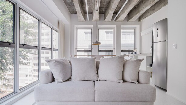 Ein modernes Wohnzimmer mit einem hellen Sofa, großen Fenstern und einer Küchenzeile mit einem Edelstahlkühlschrank.