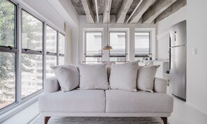 A modern living room with a light-colored sofa, large windows, and a kitchen area with a stainless steel refrigerator. - Tabas Edifício Bianca (São Paulo)